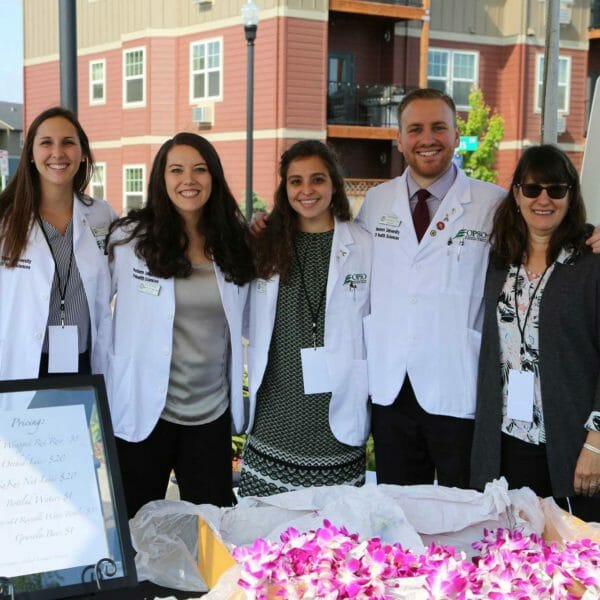Students wearing white coats and assisting at the flower/lei booth during the COMP-Northwest ceremony. There is a table in front of them with bright pink orchid leis. Students wearing white coats and assisting at the flower/lei booth during the COMP-Northwest ceremony. There is a table in front of them with bright pink orchid leis.