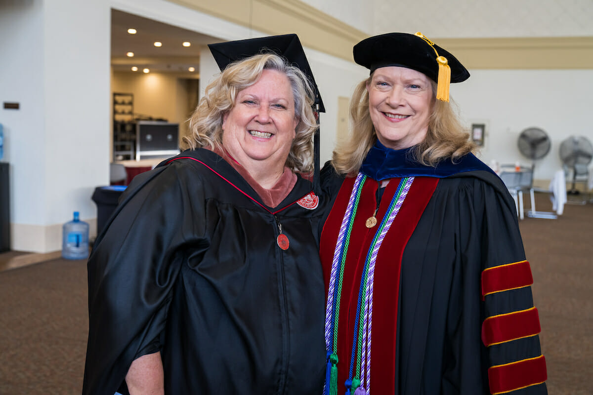 College of graduate nursing faculty member standing with the college dean. Both are wearing black robes and caps with tassels. The dean is wearing red velvet strips on their gown and blue, green, and purple and white braided honors cords around their neck. Both are wearing a zipper pull on their robe and academic hoods around their neck. College of graduate nursing faculty member standing with the college dean. Both are wearing black robes and caps with tassels. The dean is wearing red velvet strips on their gown and blue, green, and purple and white braided honors cords around their neck. Both are wearing a zipper pull on their robe and academic hoods around their neck.