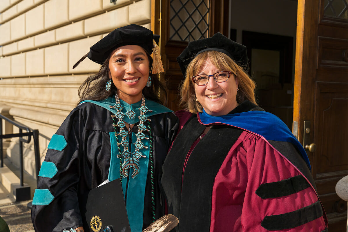 University administrator wearing burgundy regalia and a blue hood posing with an optometry graduate wearing their academic regalia with bright blue velvet. University administrator wearing burgundy regalia and a blue hood posing with an optometry graduate wearing their academic regalia with bright blue velvet.