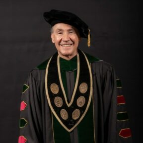 President Farias Eisner A smiling man in formal academic regalia, including a black doctoral gown with velvet panels and chevrons in red and green, a black tam with a gold tassel, and a ceremonial medallion with gold medallions hanging from a black and gold-trimmed stole. The background is a solid dark color.