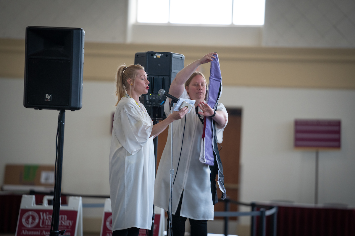 Commencement assistants demonstrating to graduates how to hold an academic hood over their arm while they walk across stage. One assistant is speaking into a microphone while the other is about to place a hood on their arm. Commencement assistants demonstrating to graduates how to hold an academic hood over their arm while they walk across stage. One assistant is speaking into a microphone while the other is about to place a hood on their arm.