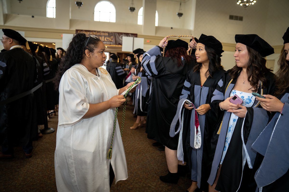 A commencement assistant holding a clipboard and green and gold honors cord is speaking to a row of graduates where black gowns with grey velvet strips on the front. A commencement assistant holding a clipboard and green and gold honors cord is speaking to a row of graduates where black gowns with grey velvet strips on the front.