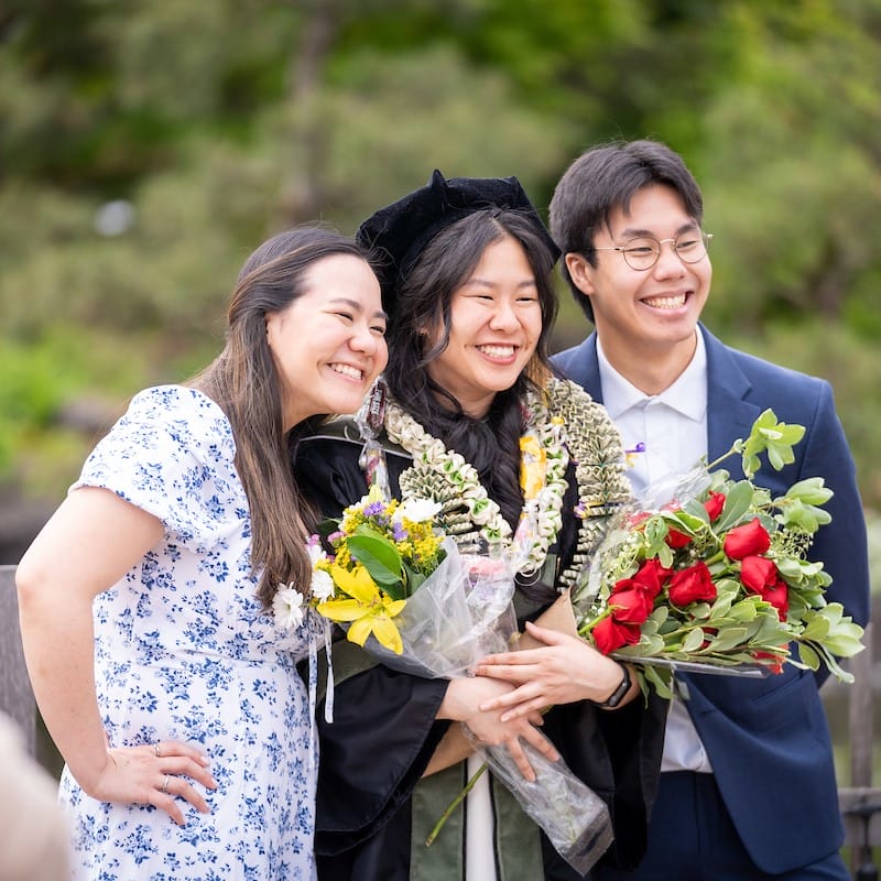 Graduate posing with their family members while holding a few large bouquets of flowers and wearing several leis.
