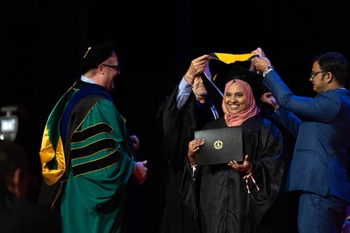 hooding-graduate This images shows a graduate smiling at the camera while her family hooders place her hood over her shoulders.
