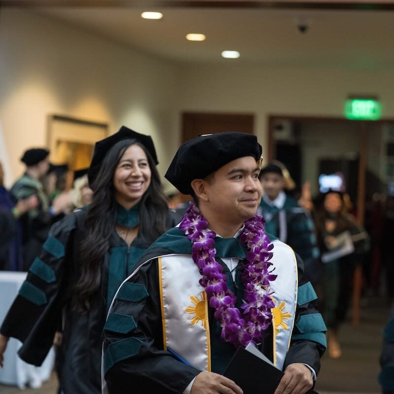 051425 CHS Commencement 168 Graduates in black and teal academic gowns and velvet caps walk in a ceremony procession indoors. The man in front wears a decorated white stole with gold sun symbols and a purple lei, holding a diploma folder, while the smiling woman behind him also wears a black and teal gown. Faculty and attendees applaud along the sides.