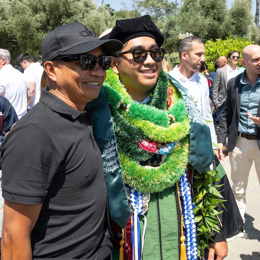 25COMP Grad A graduate wearing a green academic gown, doctoral cap, and multiple colorful leis smiles while posing for a photo with a supporter outside the commencement ceremony. The supporter is wearing a black polo shirt, sunglasses, and a cap. Other attendees and greenery are visible in the background.
