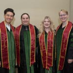 051225 WesternU Oregon Commencement 036 Four smiling graduates pose indoors, each wearing a black gown with green accents and a long, maroon and gold academic stole. The stoles are embroidered with text including "WESTERN U" and "OMM FELLOW" and the graduates' names.
