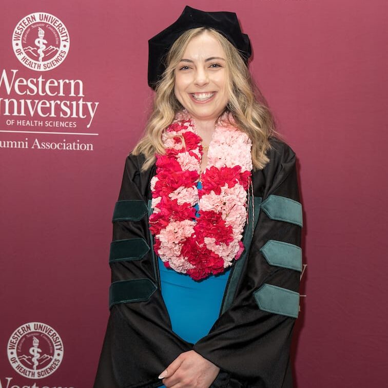 051225 WesternU Oregon Commencement 040 A smiling female graduate with blonde hair stands in front of a maroon backdrop featuring the Western University logo. She is wearing a black and teal academic gown, a black velvet cap, and thick pink and red flower leis around her neck over a blue dress.