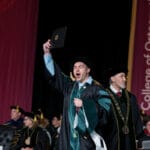 051225 WesternU Oregon Commencement 338 A graduate in a black gown and green academic hood enthusiastically raises a diploma folder high above his head on stage, mouth open in celebration. An official in black academic robes and a chain of office stands next to him. The background includes a large maroon banner that partially reads "College of Osteopathy of the Pacific-Northwest."