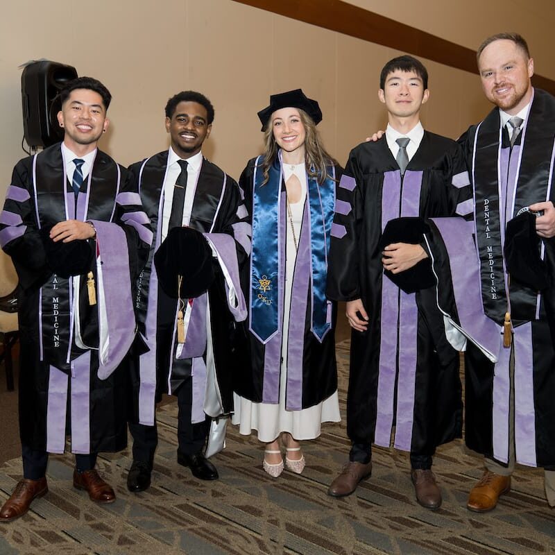 051325 WesternU CDM Commencement 005 ive graduates in black and lavender doctoral regalia stand indoors posing for a photo. Four wear black gowns with purple velvet panels and hold their graduation caps, while one in the middle wears a similar gown plus a blue honor stole and her cap on. They are smiling and standing in a row in a banquet-style room.
