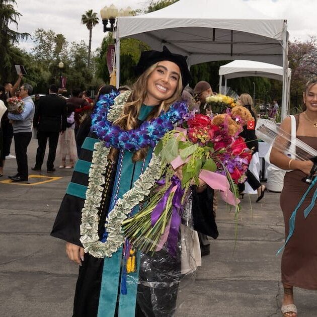 051325 WesternU CO Commencement 188 A smiling graduate in a black and teal academic gown and velvet cap stands outdoors, holding a large bouquet of flowers and wearing multiple colorful leis, including one made of currency. She is posing with a group of three younger women who are also smiling, in a sunny outdoor plaza with palm trees and white tents.