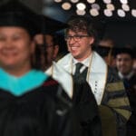 051325 WesternU COP Commencement 054 A graduate with curly hair and glasses, wearing a black, gold, and white academic gown and a stole that reads "GRADUATE," smiles broadly while walking among a crowd of other graduates. The background is dark with blurred bokeh lights.
