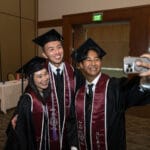 051425 CGN Commencement 006 Three graduates in black cap and gowns, and maroon and white stoles pose for a selfie in a reception hall. All three are smiling, and the graduate on the right is holding a phone to take the picture.