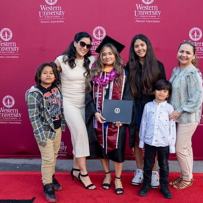 051425 CGN Commencement 256 A graduate in a black cap and gown, maroon stole, and flower leis stands on a red carpet with five family members (two women and three young boys). They are posing in front of a tall, red backdrop with the Western University Alumni Association logo and a vertical banner to the right that reads "CONGRATULATIONS GRADUATES."