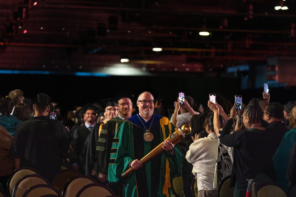 051425 CHS Commencement 023 The University Marshal holding the mace and leading the processional down the aisle to the stage. Several family members can be seen in the photo holding their cell phones high to capture the moment.