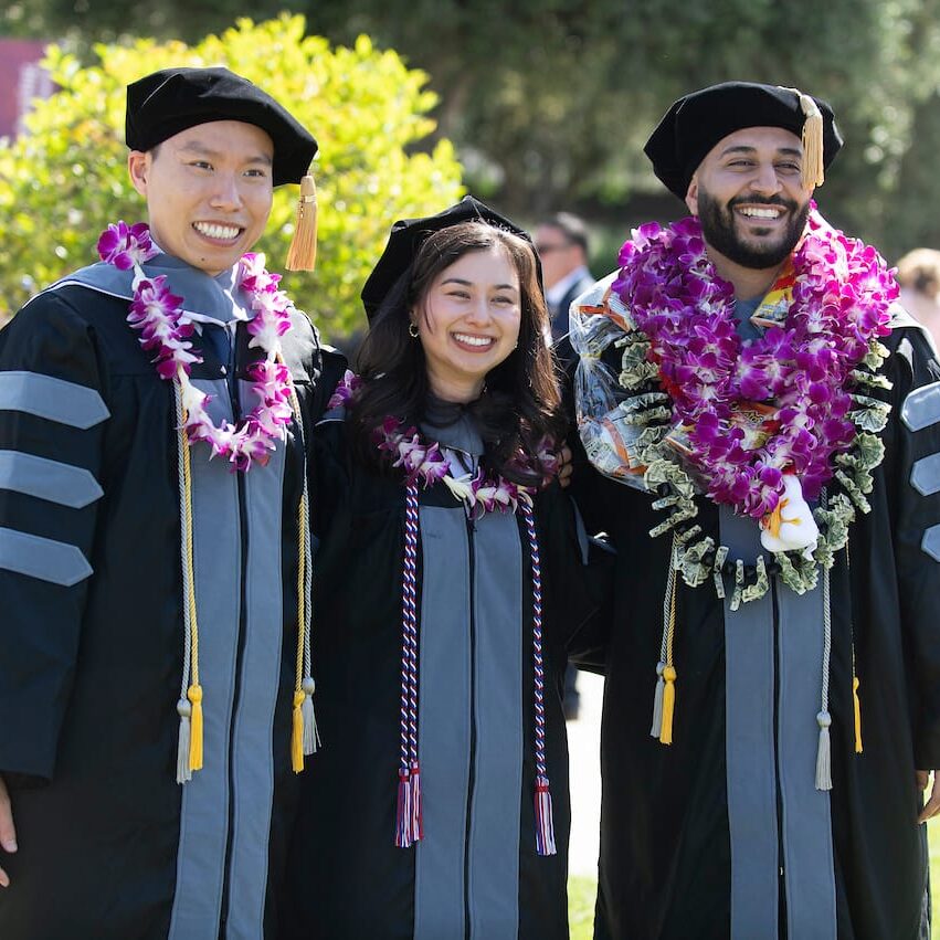 25VET Grad Three graduates in black and gray academic regalia and black velvet caps pose together outdoors on a sunny day. Each wears purple and white flower leis around their necks. They are all smiling.