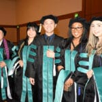 50407720-00701-0028 A group of six smiling graduates, standing in a line indoors. They are all wearing black academic gowns, black velvet caps, and teal and black academic hoods. The second graduate from the left wears a purple and white stole.