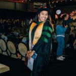 WU CPM 2025 Graduation A smiling female graduate in a black gown, green academic hood, and a gold stole that reads "Podiatric Medicine" stands in the aisle of a dark commencement venue. She is holding a program and diploma case, with rows of chairs and other people visible in the background.