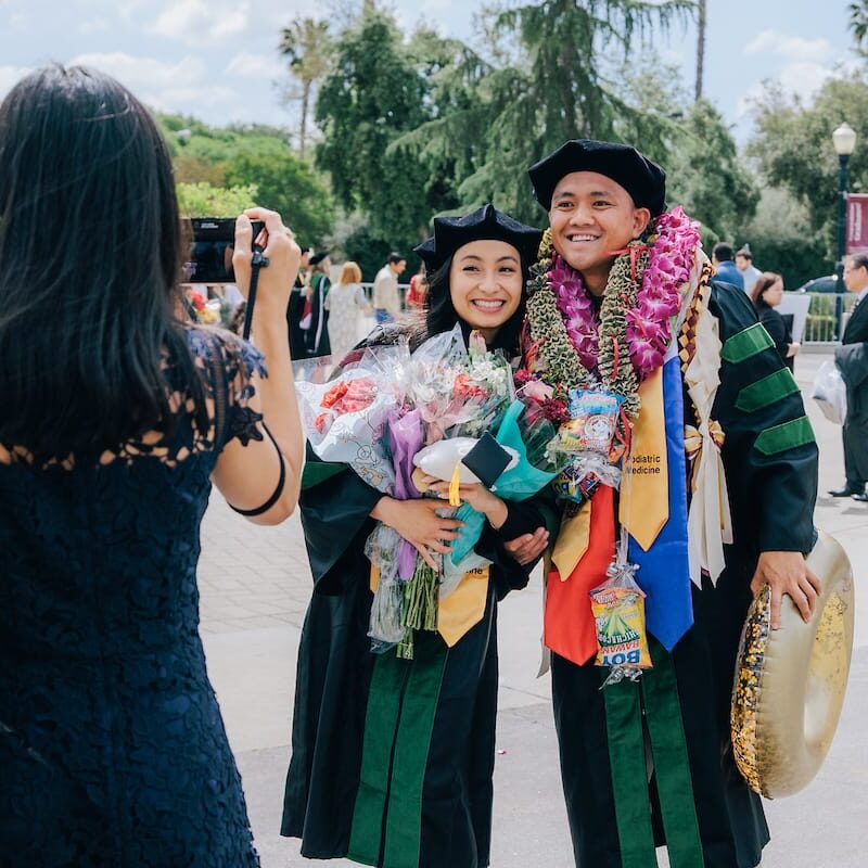 WU CPM 2025 Graduation Two graduates in black gowns with green velvet panels and doctoral caps stand outdoors smiling and holding bouquets. They are covered in colorful leis and ribbons. A woman in a navy lace dress stands in front of them taking their photo. Trees and people are visible in the background on a sunny day.