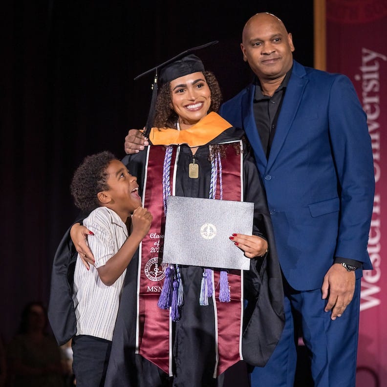 sq Image of a graduate posing with a man in a blue suit and a child wearing a white shirt. The child is looking up and excitedly smiling at the graduate.